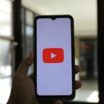 A hand holding a smartphone displaying the YouTube app indoors.